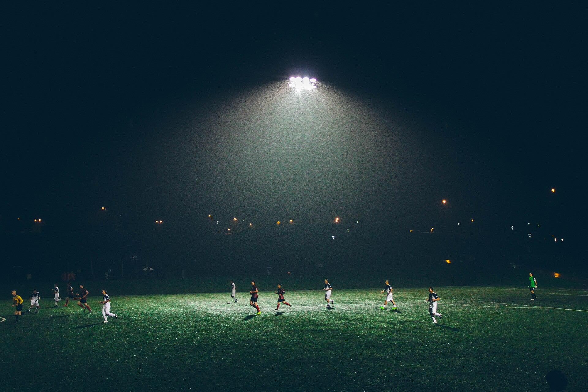 Adult players running across a floodlit soccer pitch at night